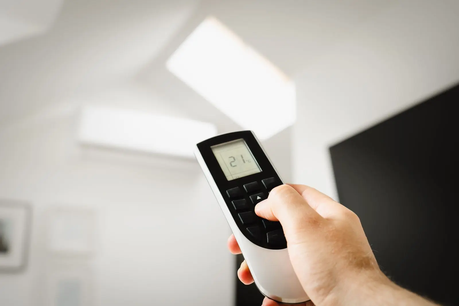 Hand holding a remote pointed at a white wall-mounted split-system air conditioner in a bright modern room
