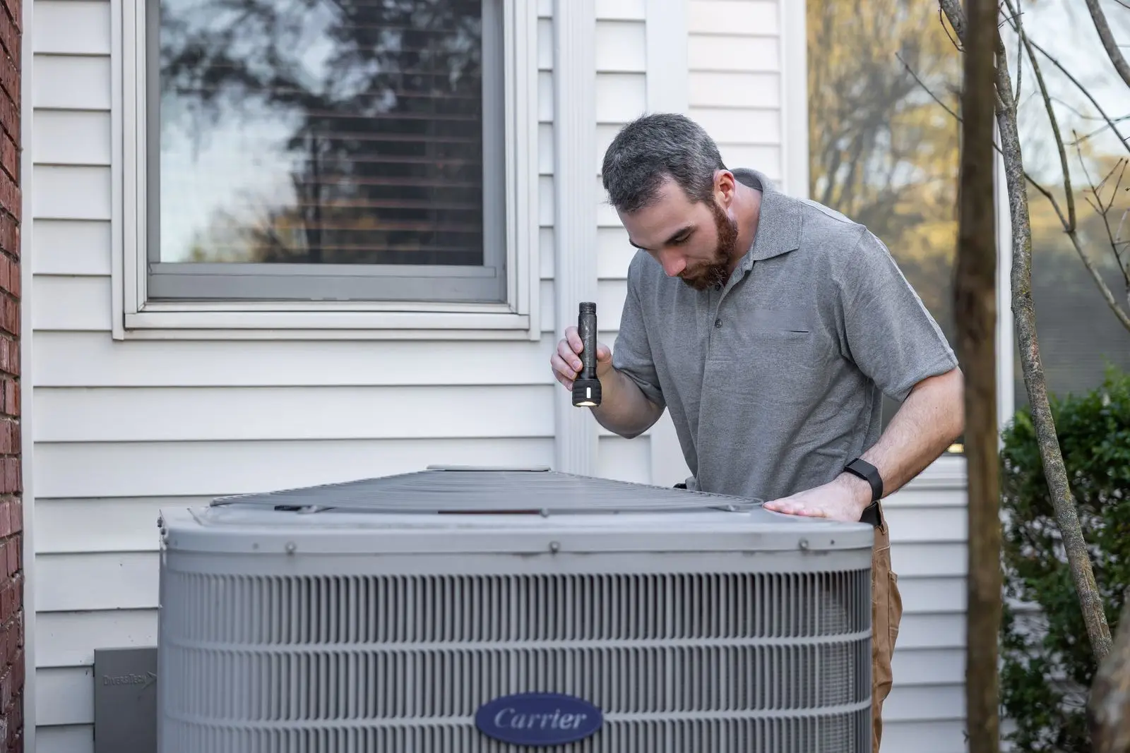 HVAC technician inspecting an outdoor air-conditioning condenser unit beside a house