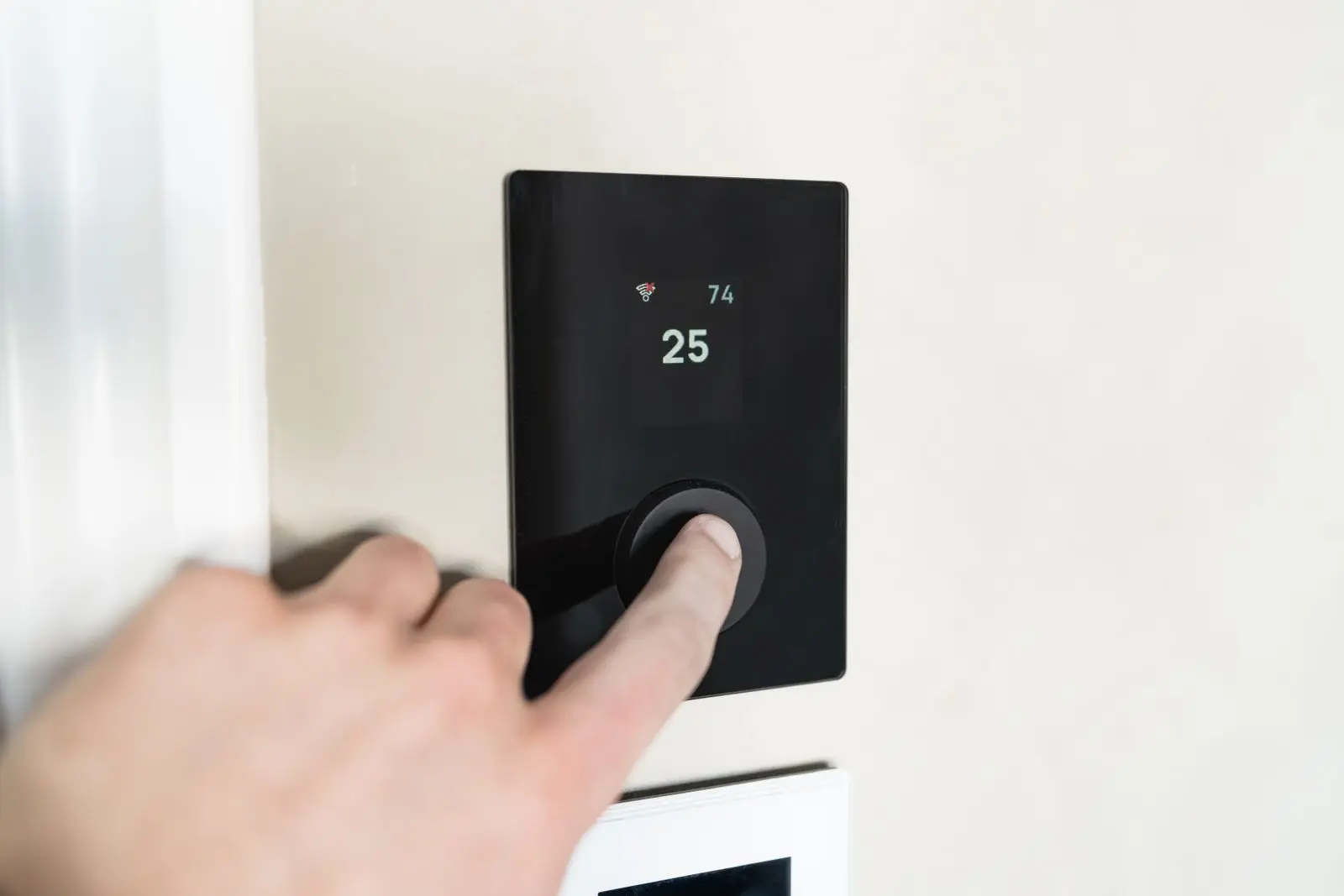 Finger adjusting a modern wall-mounted smart thermostat set to 25 degrees
